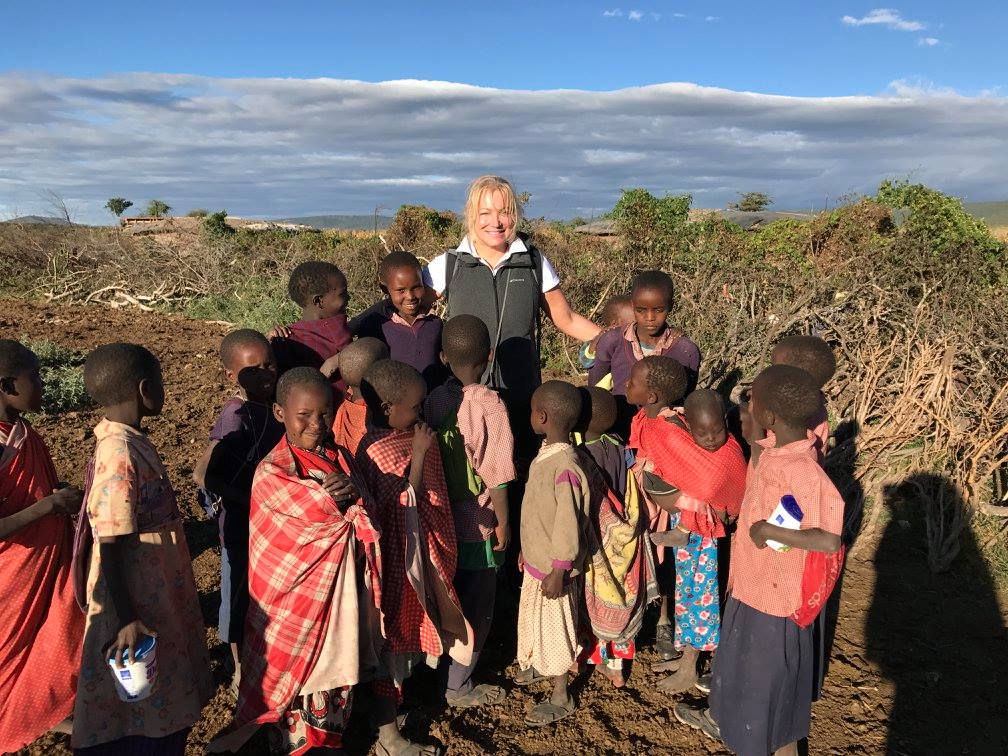A person standing outdoors with a group of children, under a cloudy sky. The children are wearing various colorful clothing.