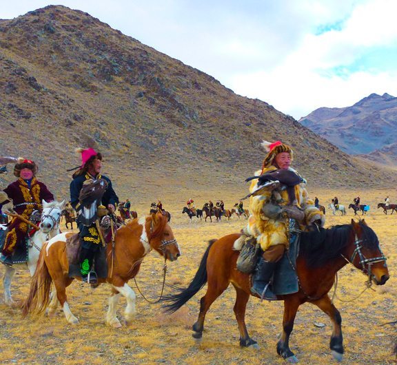 People on horseback wearing traditional attire, holding eagles in a vast, mountainous landscape.