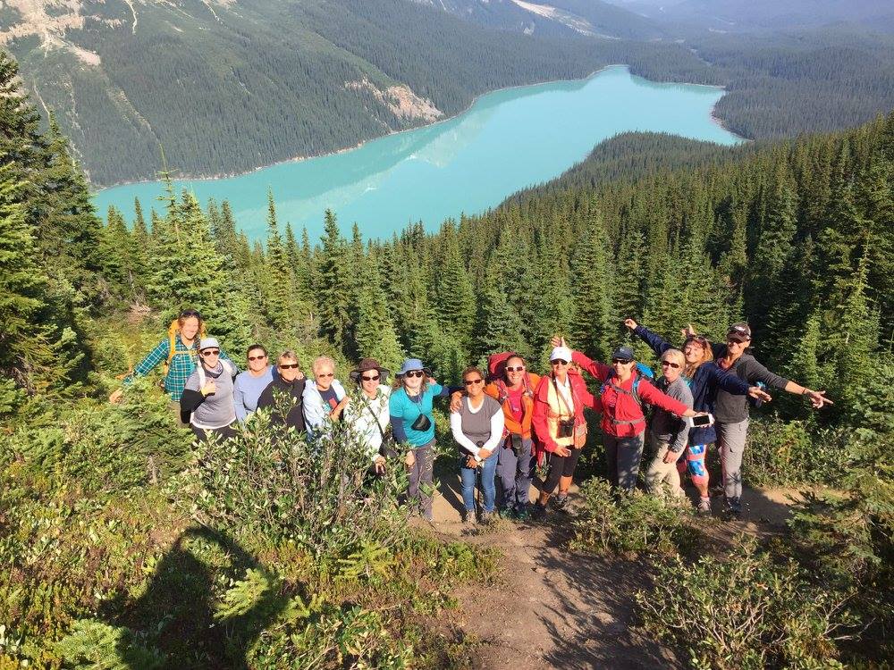 A group of people posing on a hiking trail with a turquoise lake and forested mountains in the background.