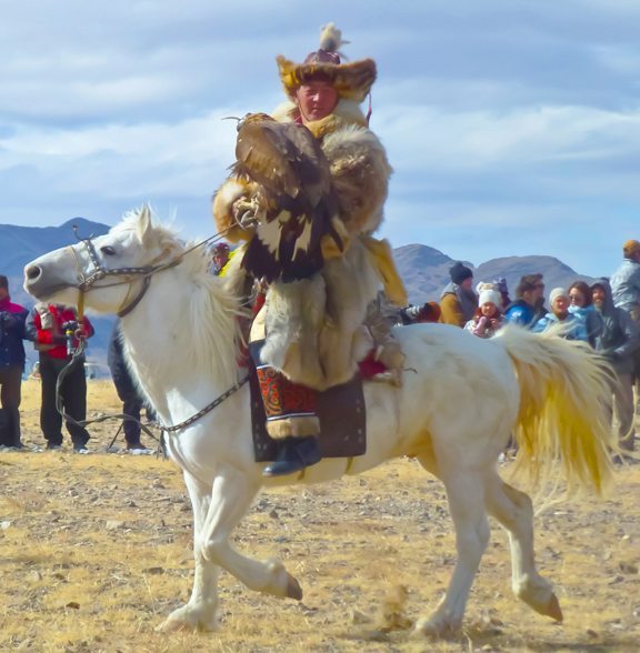 A person in traditional attire rides a white horse while holding a large bird, with a crowd and mountainous landscape in the background.