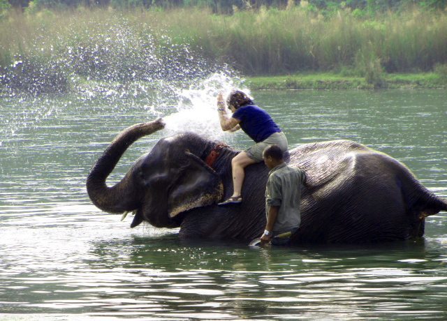 Person sitting on an elephant in the water, splashing with its trunk, while another person stands nearby.