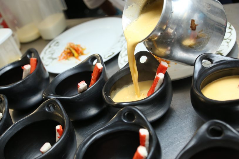 Creamy yellow soup is being poured from a metal pot into black bowls lined up in a row, each containing a breadstick.