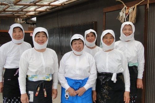 Six women wearing traditional white clothing and head coverings stand together, smiling, in an outdoor setting.