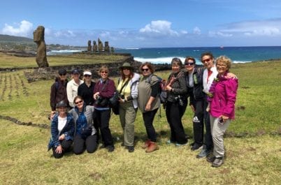 A group of people pose for a photo on grassy terrain near several stone statues with the ocean in the background.
