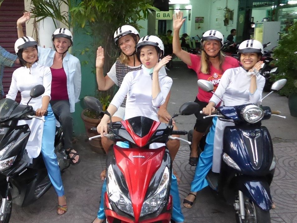 A group of people wearing helmets on scooters, smiling and waving at the camera.