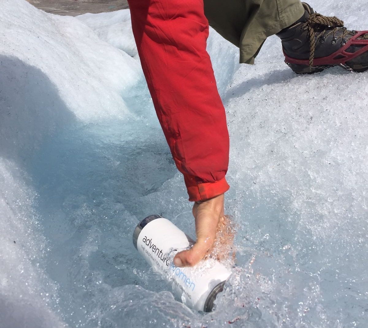 A person in a red jacket and hiking boots places a white water bottle in a stream on icy terrain.
