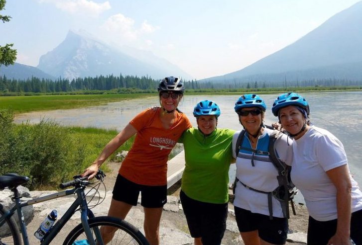 Four people wearing helmets stand with bicycles by a lake, surrounded by mountains and trees on a clear day.