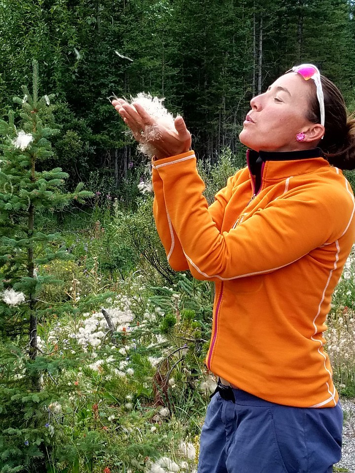 A person in an orange jacket blows white seeds from their hands in a forested area.