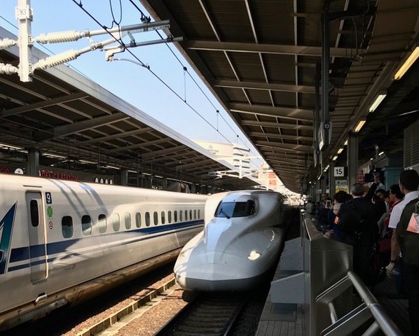 Two bullet trains are stopped at a train station platform. Passengers stand nearby, waiting to board. Overhead cables and station roofing are visible.