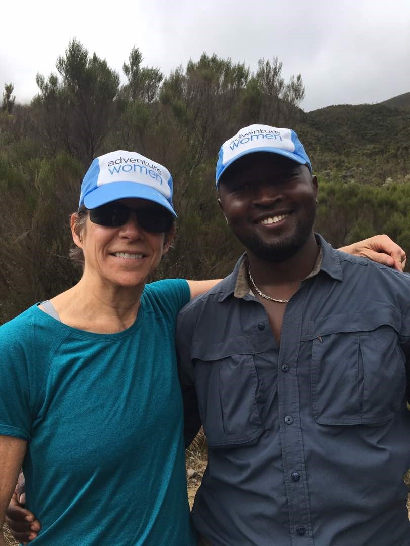 Two people wearing blue "adventure women" hats and casual outdoor clothing pose together, smiling, in a natural setting with green foliage in the background.