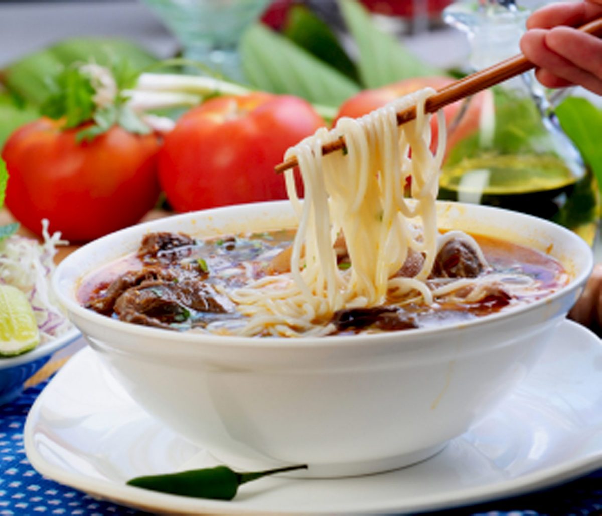 A bowl of noodle soup with meat and vegetables, with chopsticks lifting noodles. Fresh tomatoes and greens in the background.