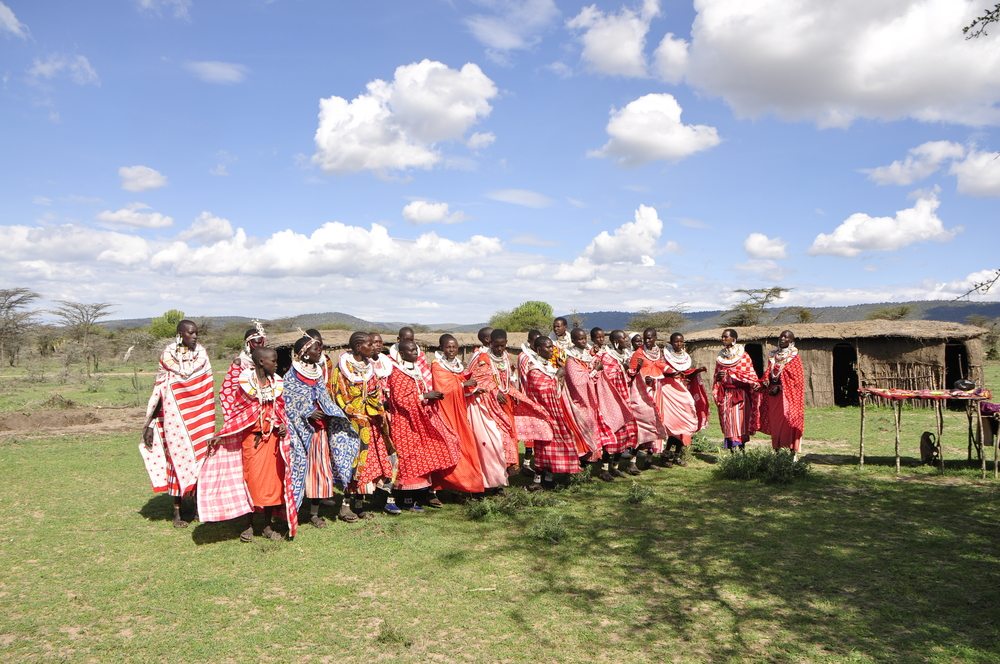 A group of people in colorful traditional attire dance outdoors on a grassy field under a partly cloudy sky. A thatched hut and a table are visible in the background.
