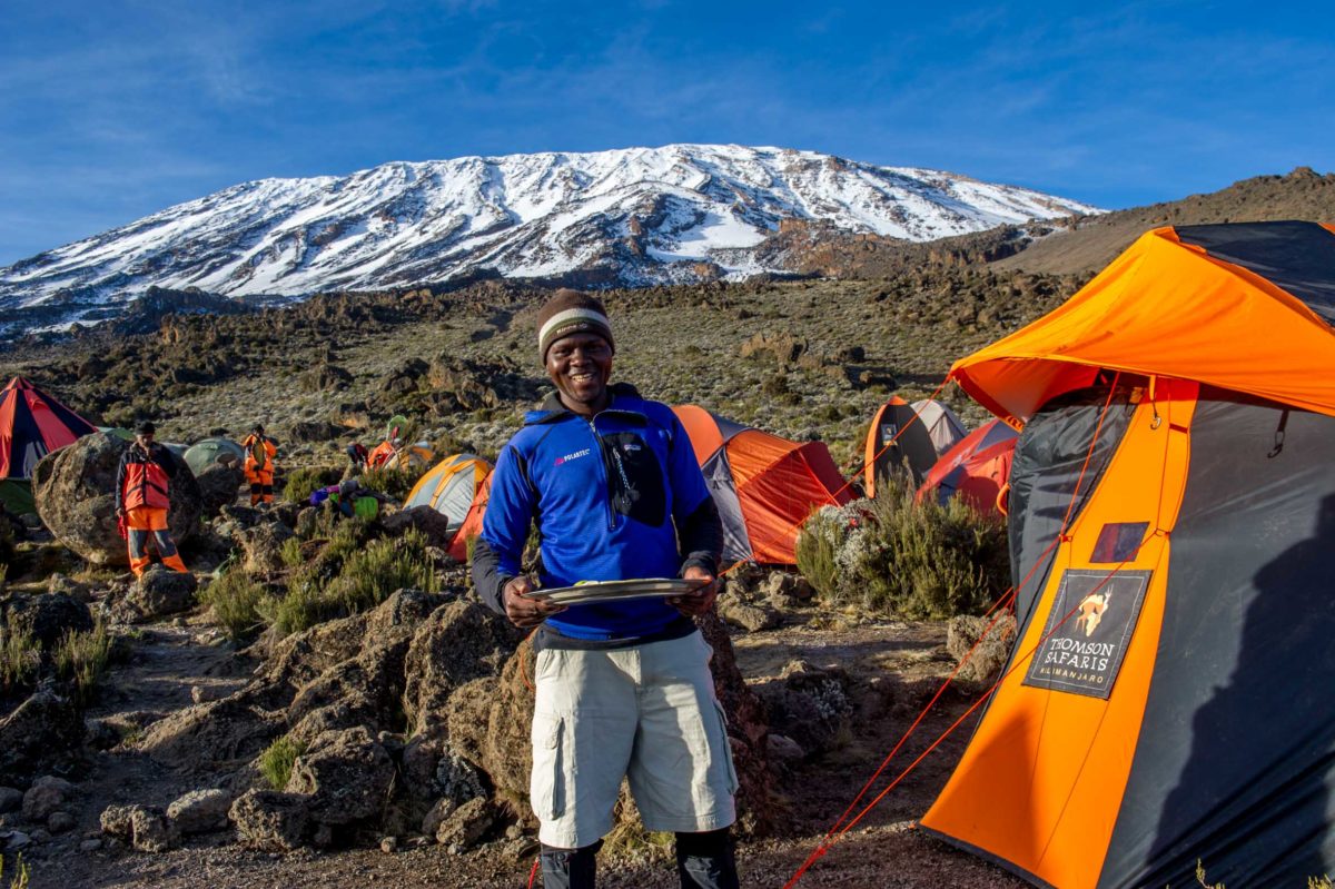A man stands smiling near a campsite with colorful tents, set against a backdrop of a snow-capped mountain.