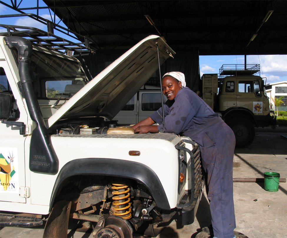 A mechanic wearing a headscarf works on the engine of a white vehicle inside a garage. Another vehicle is visible in the background.