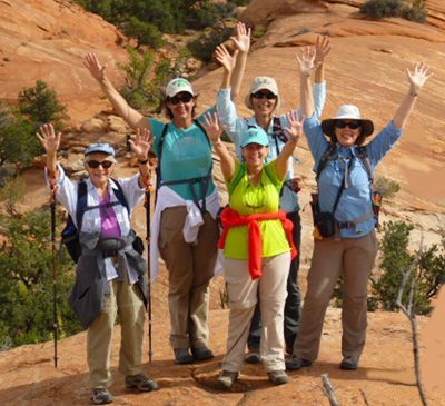 Five people on a rocky trail, raising arms and smiling, with hiking gear and walking sticks. Desert landscape in the background.