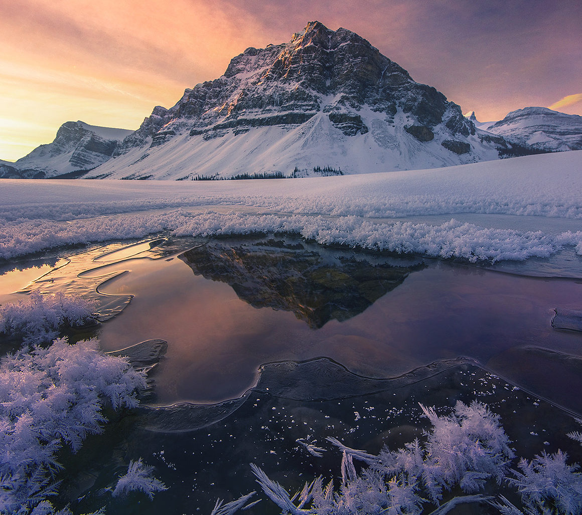 Snow-covered mountain under a colorful sky is reflected in a partially frozen lake with ice crystals along the edges.