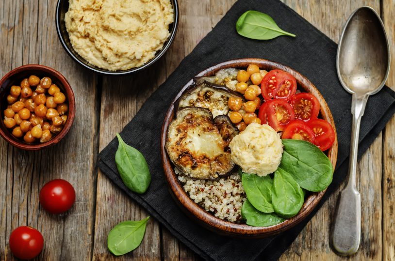 A wooden bowl with quinoa, roasted eggplant, cherry tomatoes, chickpeas, spinach, and a dollop of hummus. A spoon, tomatoes, and more chickpeas are nearby on a wooden table.