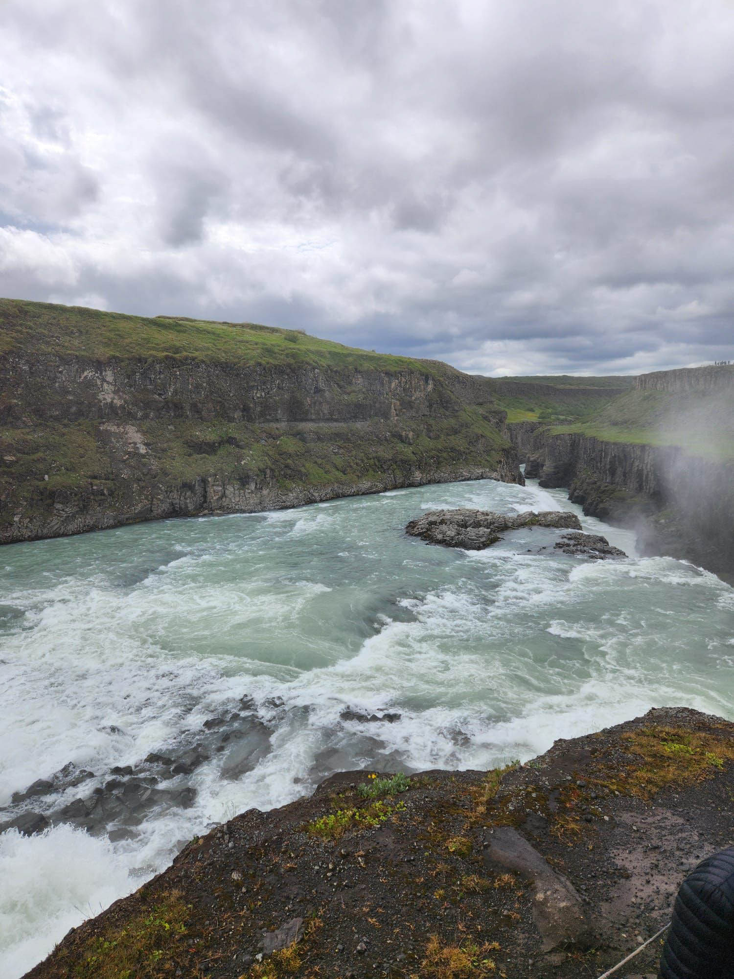 Wide river with rapids flowing through a rocky canyon, surrounded by grassy cliffs under a cloudy sky.