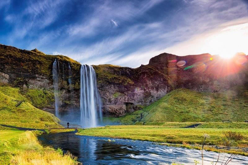 A tall waterfall cascades down a rocky cliff into a river, surrounded by green hills. The sun is setting, casting a bright glow in the sky.