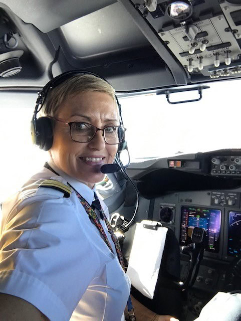 A pilot with short hair and glasses is smiling while seated in an aircraft cockpit, wearing a headset and uniform.
