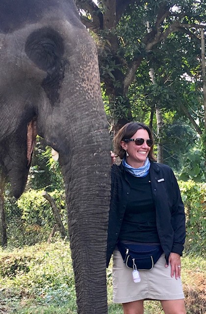 A person stands beside an elephant, smiling, in an outdoor setting with trees in the background.