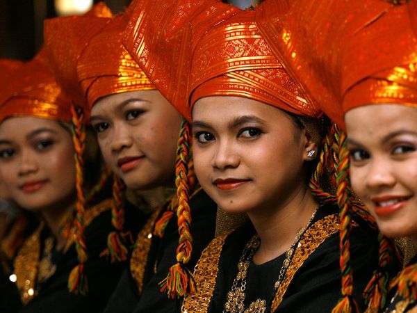 Four women wearing traditional orange headgear and black attire with gold accents, looking towards the camera.