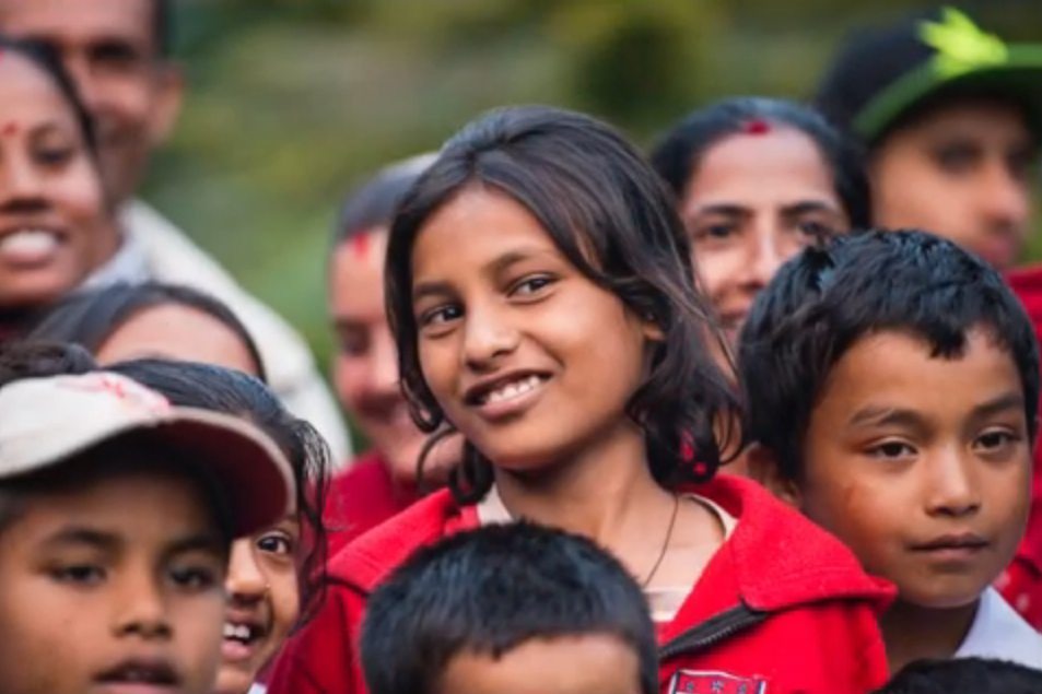 A group of smiling children and adults, some wearing red jackets, gather closely for a photo.