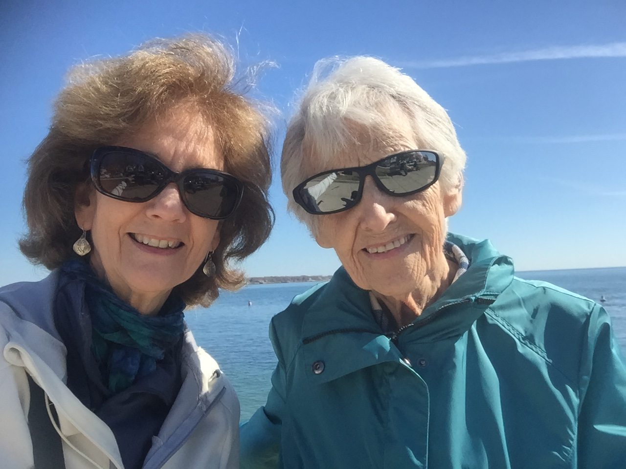 Two women wearing sunglasses pose for a selfie by the water on a sunny day.