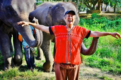 A person in a red shirt smiles with outstretched arms as two elephants stand beside them, one with its trunk around their neck, in a grassy area with trees in the background.