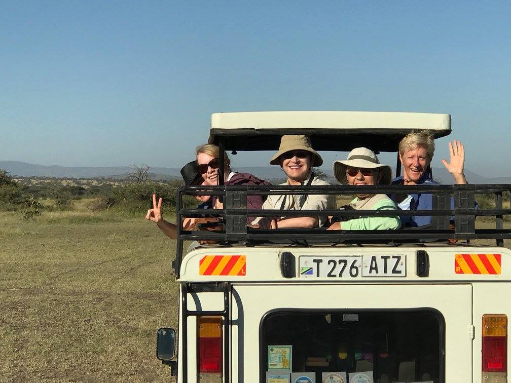 Four people sitting in an open safari vehicle, smiling and waving, with a grassy landscape in the background.