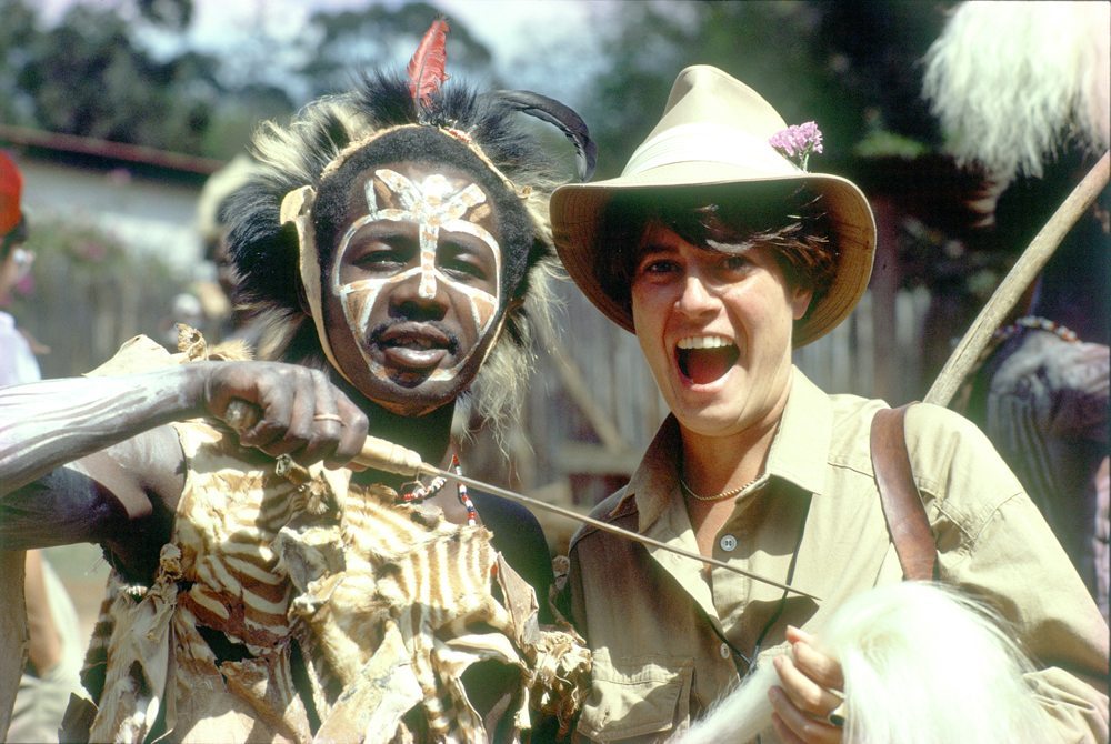 Person in traditional attire and face paint stands beside another person wearing a safari hat, both smiling at the camera, outdoors.