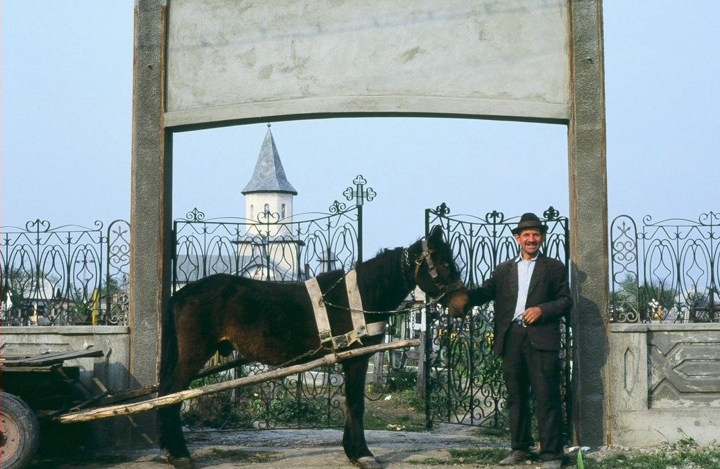 A man stands beside a horse-drawn cart under an archway, with a church steeple visible in the background.