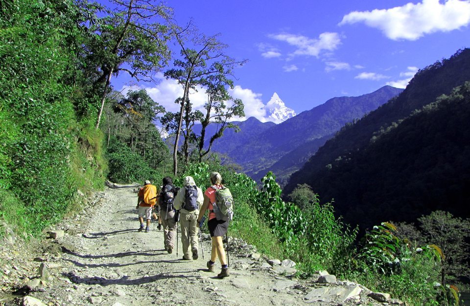 Hikers walking along a rugged mountain path surrounded by lush greenery and distant peaks under a bright blue sky.