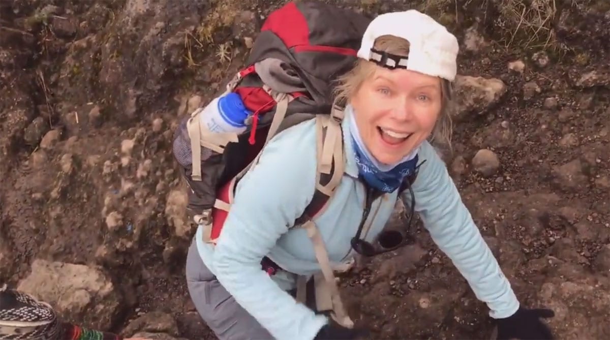 A person in outdoor gear climbs a rocky terrain, smiling at the camera. They wear a backpack, cap, and a blue shirt.