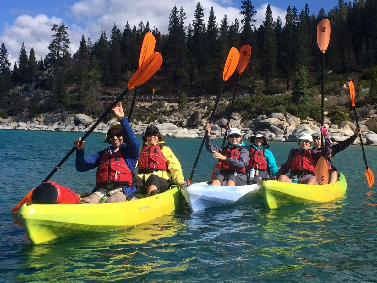 Group of five people kayaking on a clear lake surrounded by trees and rocks, holding orange paddles above their heads.