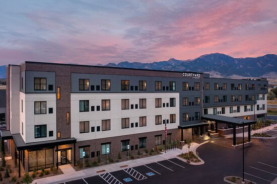 A modern hotel building with a mix of dark and light exterior under a pinkish sky, featuring a mountain range in the background and an adjacent parking lot.