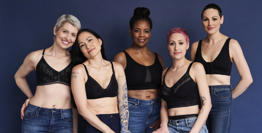 Five women in black bras and jeans stand against a dark blue background, smiling at the camera.
