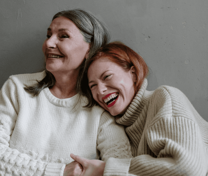 Two women in sweaters, one with gray hair and the other with red hair, smiling and leaning against each other against a plain wall.