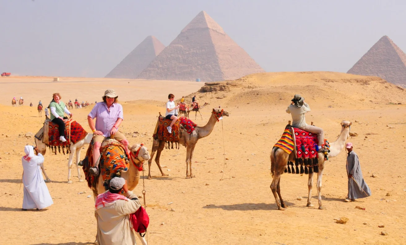 Tourists ride camels near the Pyramids of Giza, guided by locals in traditional clothing, with a clear sky overhead.