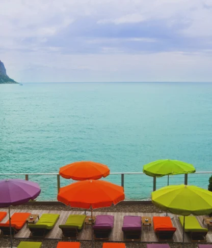 Colorful umbrellas and lounge chairs line a pebble beach overlooking a calm turquoise sea, with a rocky cliff and cloudy sky in the background.