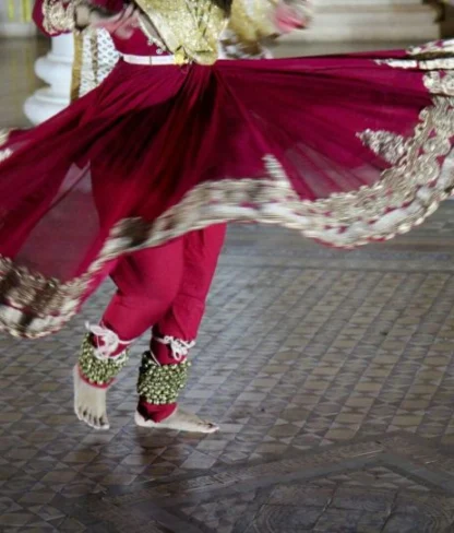 A person dances, wearing a red traditional outfit with gold embroidery, twirling on a patterned floor.