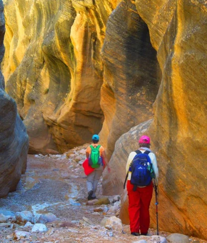 Two hikers wearing backpacks and hats walk through a narrow, rocky canyon with sunlight illuminating the stone walls.
