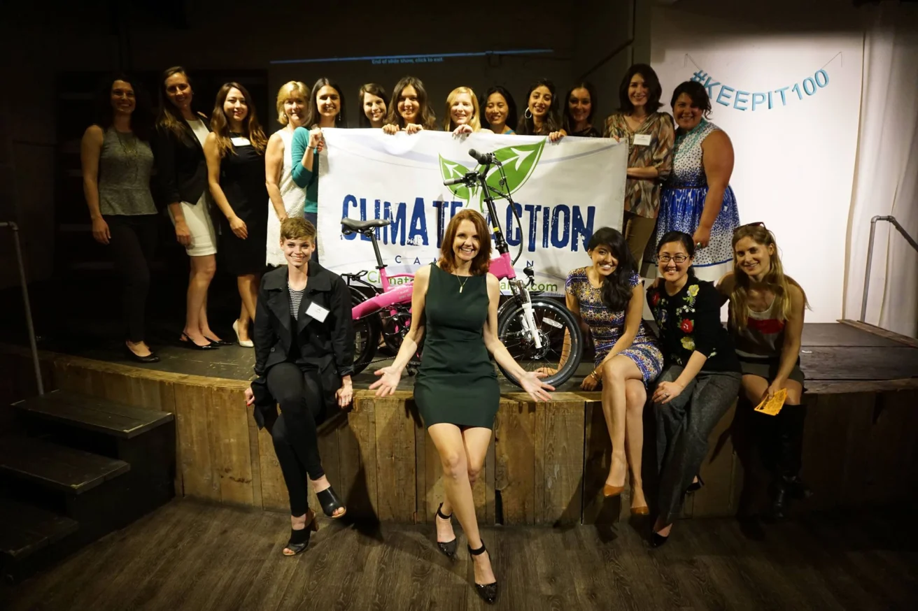 A group of smiling people pose with a "Climate Action" banner and a bicycle on a stage indoors.