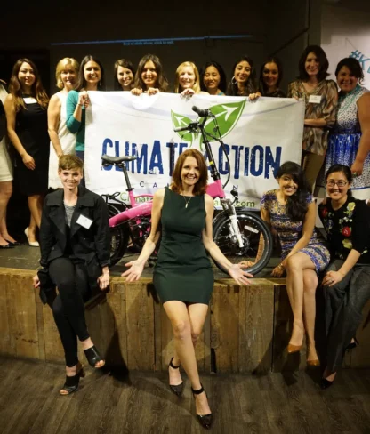 A group of smiling people pose with a "Climate Action" banner and a bicycle on a stage indoors.