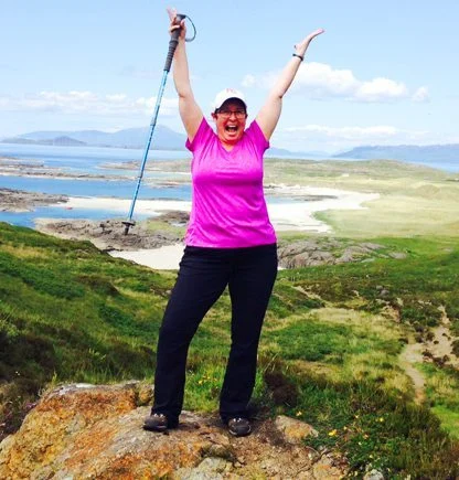 Person standing on a rock with arms raised, holding a walking stick, overlooking a green landscape and coastline under a clear blue sky.