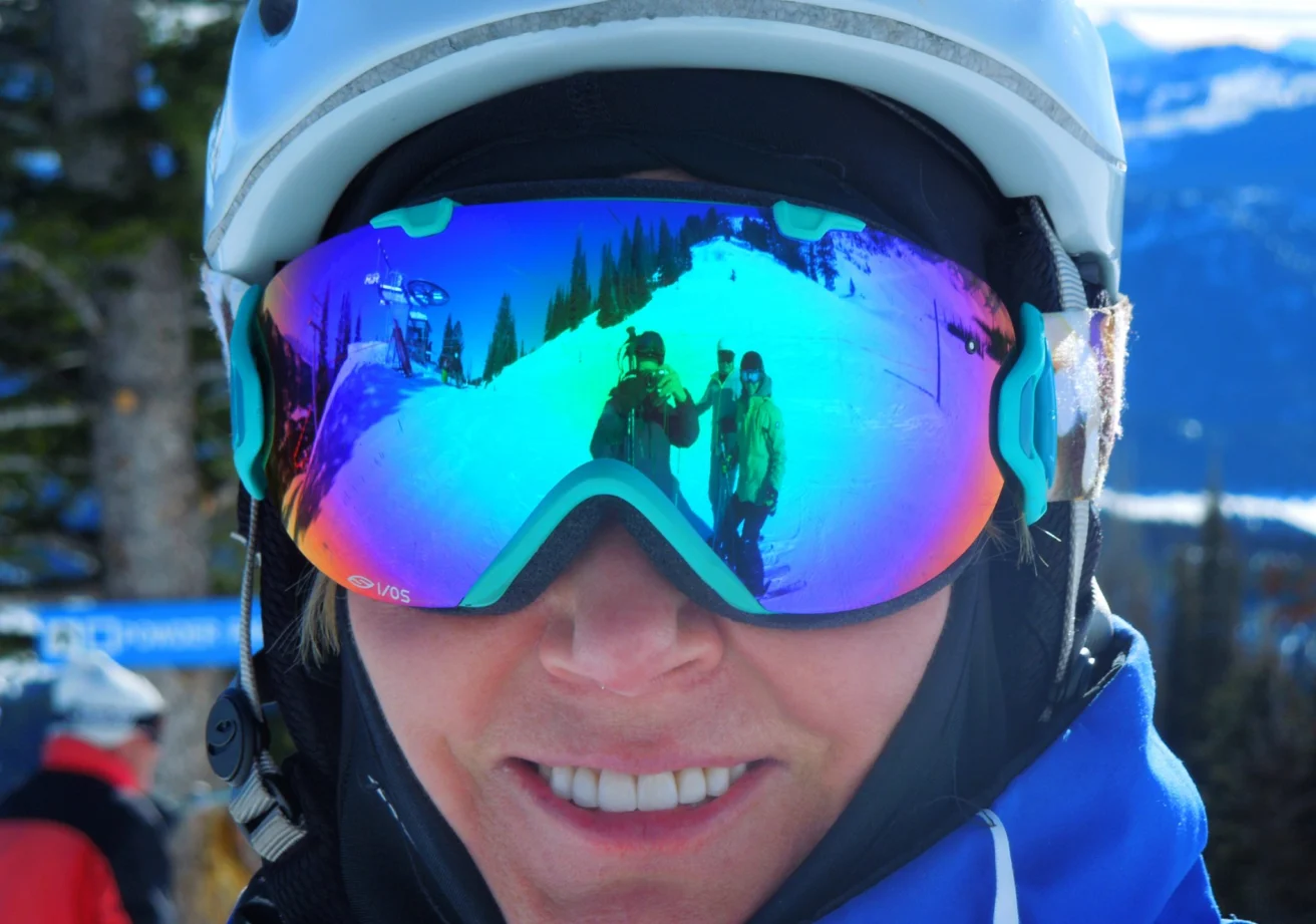 A person wearing mirrored ski goggles captures a snowy mountain reflection with another person waving. They are dressed in winter gear, including a helmet.