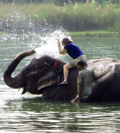 Person sitting on an elephant in the water, splashing with its trunk, while another person stands nearby.