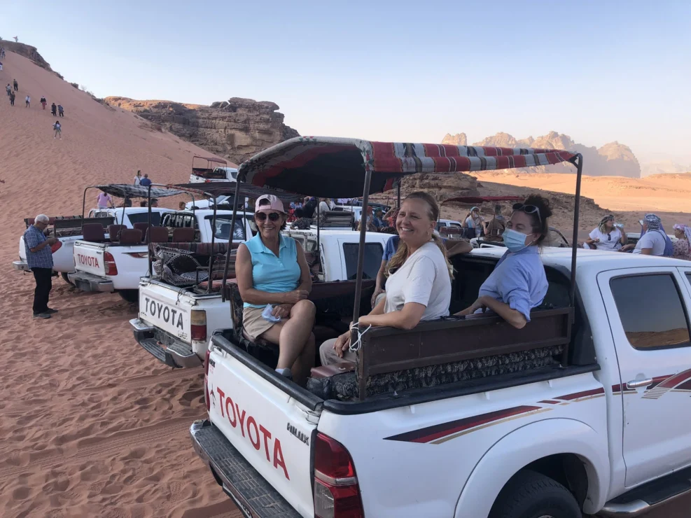 People sitting in the back of parked pickup trucks on desert terrain with rocky formations in the background.