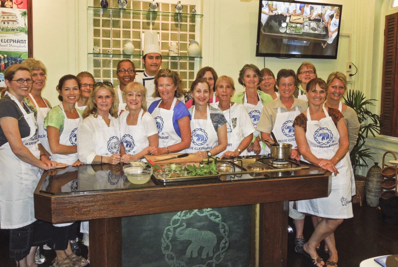 A group of people wearing aprons pose in a kitchen. A chef stands in the back. There are ingredients and cooking tools on the counter.