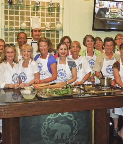 A group of people wearing aprons pose in a kitchen. A chef stands in the back. There are ingredients and cooking tools on the counter.